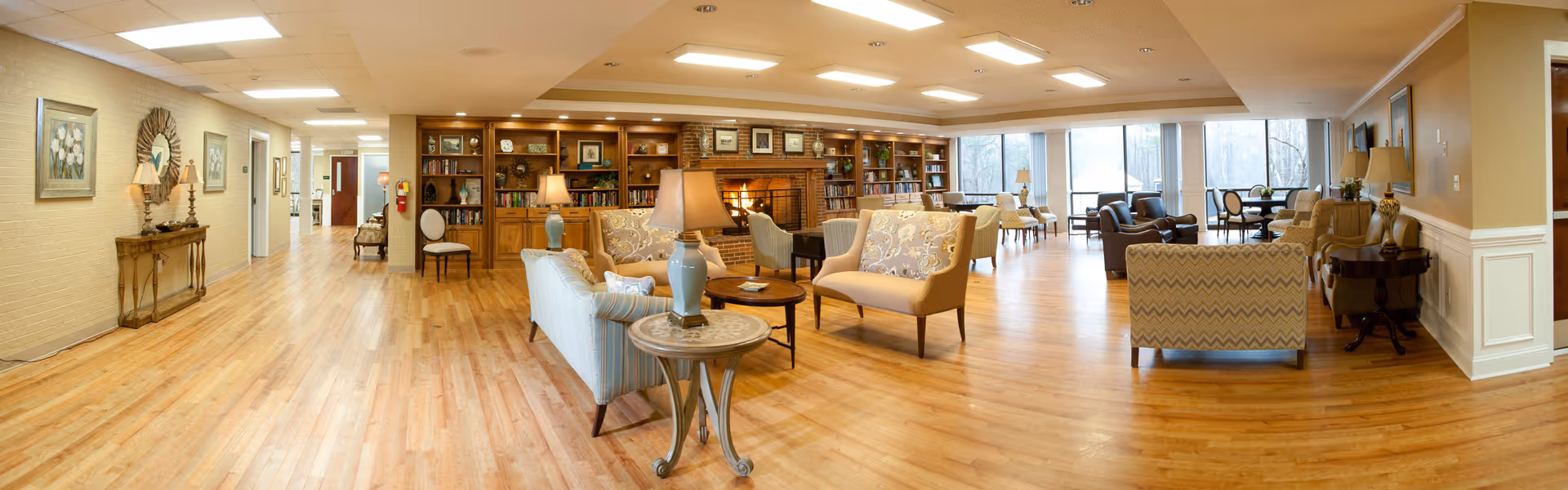 A spacious and well-lit senior living facility common area with wooden floors, comfortable upholstered chairs and sofas arranged around small tables, a brick fireplace with a wooden mantel, built-in bookshelves filled with books and decorative items, large windows letting in natural light, and various lamps and framed artwork on the walls.