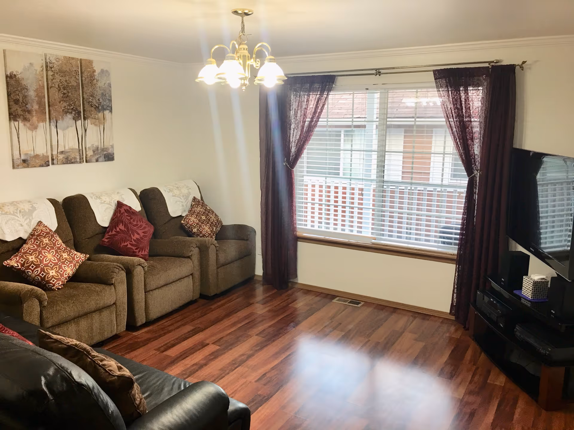A cozy living room with three brown recliner chairs adorned with patterned pillows and white doilies on the headrests. There is a black leather couch partially visible in the foreground. The room features wood flooring, a large window with white blinds and dark purple curtains, a wall-mounted flat-screen TV on the right, and a chandelier with multiple lights hanging from the ceiling. A three-panel painting of trees hangs on the wall above the recliners.