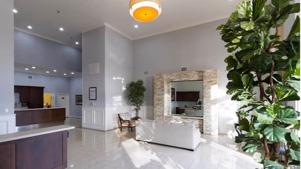 Bright modern lobby with high ceilings, a seating area in front of a stone-framed pass-through, reception desks, and large potted plants.