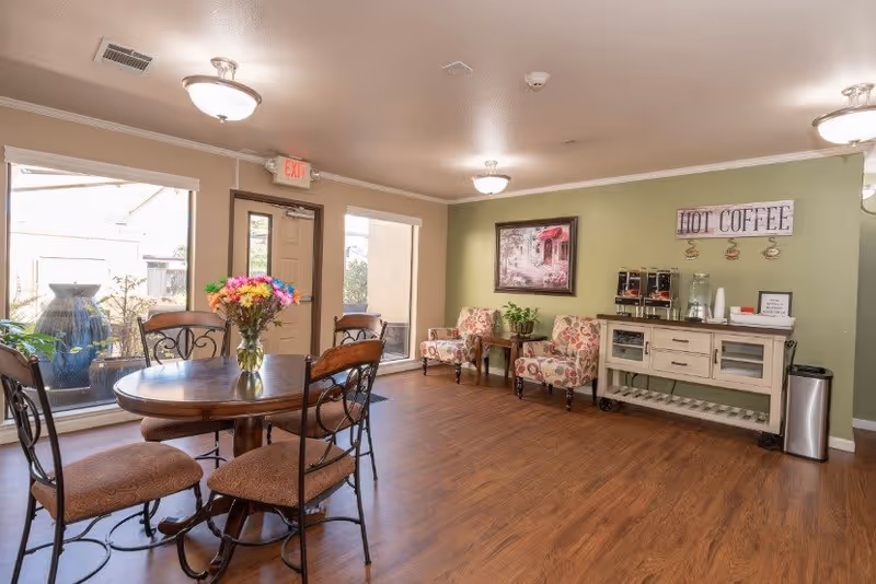 A cozy senior living common area with a round wooden table and four chairs, a vase with colorful flowers on the table, two floral upholstered armchairs with a small wooden side table between them, a coffee station with dispensers, cups, and a sign that reads 'HOT COFFEE' on the wall, large windows letting in natural light, and wood flooring.