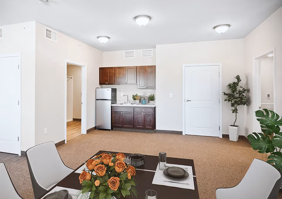 Interior view of a senior living facility apartment showing a small kitchen area with dark wood cabinets, a refrigerator, and a sink. In the foreground, there is a dining table set with plates, glasses, and a centerpiece of orange roses. The room has beige walls, carpeted floor, and two white doors. There are also two potted plants placed near the walls.