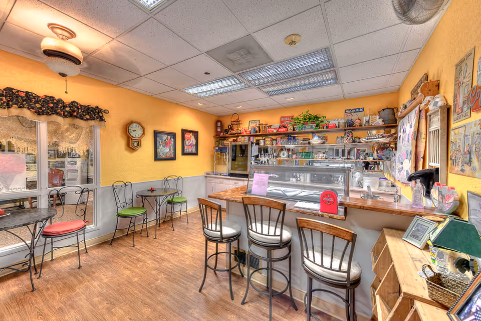 Cozy small dining area with yellow walls, wooden floor, and a counter with three bar stools. There are two small round tables with metal chairs, some with red and green cushions. The counter has a glass display case and shelves behind it filled with various decorative items, plants, and vintage signs. The room has a ceiling fan and fluorescent lights, with framed pictures and a clock on the walls.