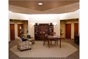 Interior common area of a senior living facility with a patterned armchair, a wooden table with chairs, a bookshelf filled with books, and several doorways leading to other rooms. The ceiling has recessed lighting and a decorative wooden panel.