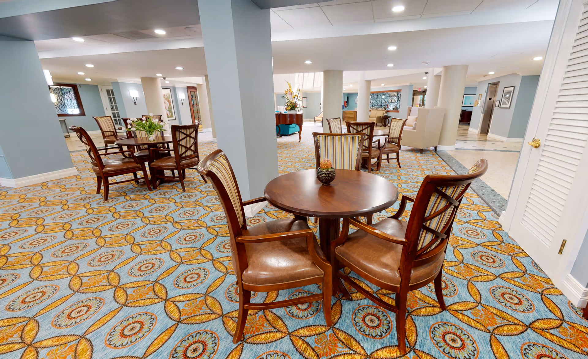 Spacious senior living common area with round wooden tables and chairs on a patterned carpet.