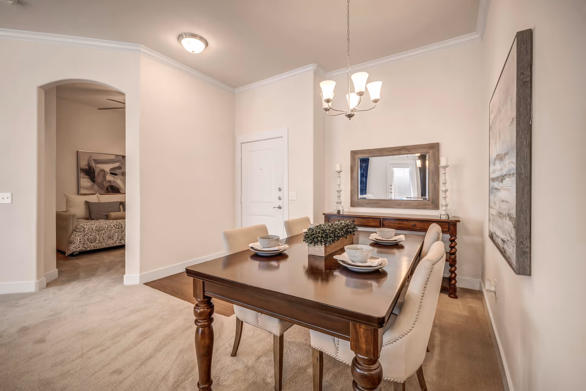 A dining room with a wooden table set for four with bowls and plates. Four beige upholstered chairs surround the table. A rectangular mirror hangs on the wall above a wooden sideboard with two candlesticks. A chandelier with five lights hangs above the table. There is a doorway leading to a bedroom with a bed and abstract artwork visible.