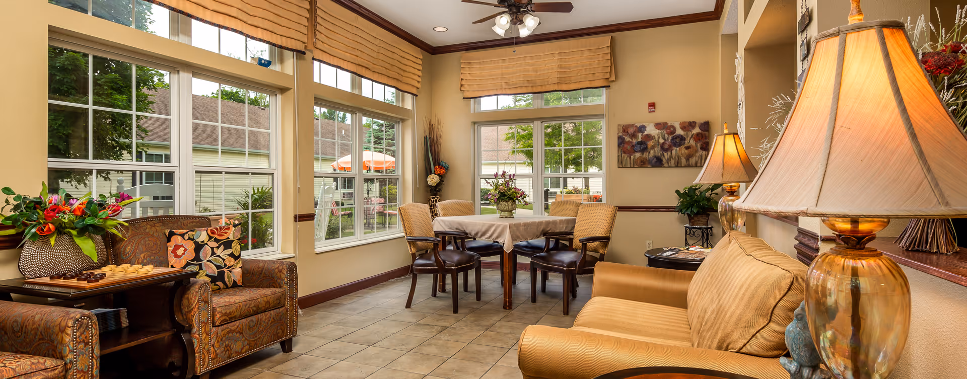 A cozy and well-lit living room area in a senior living facility with large windows showing an outdoor patio. The room features comfortable armchairs with patterned cushions, a small table with a checkers game and a flower arrangement, a dining table with four chairs, a beige sofa, and two amber-colored table lamps. The walls are decorated with floral artwork and the windows have beige roman shades.