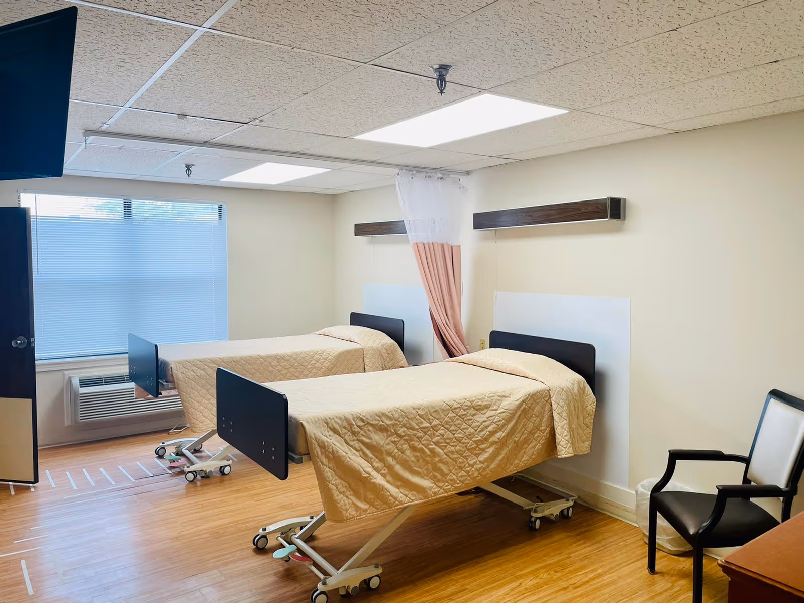 A room with two hospital-style beds covered with beige quilts, a black chair, a window with blinds, and a curtain divider between the beds. The room has a light-colored ceiling with fluorescent lights and wood-patterned flooring.
