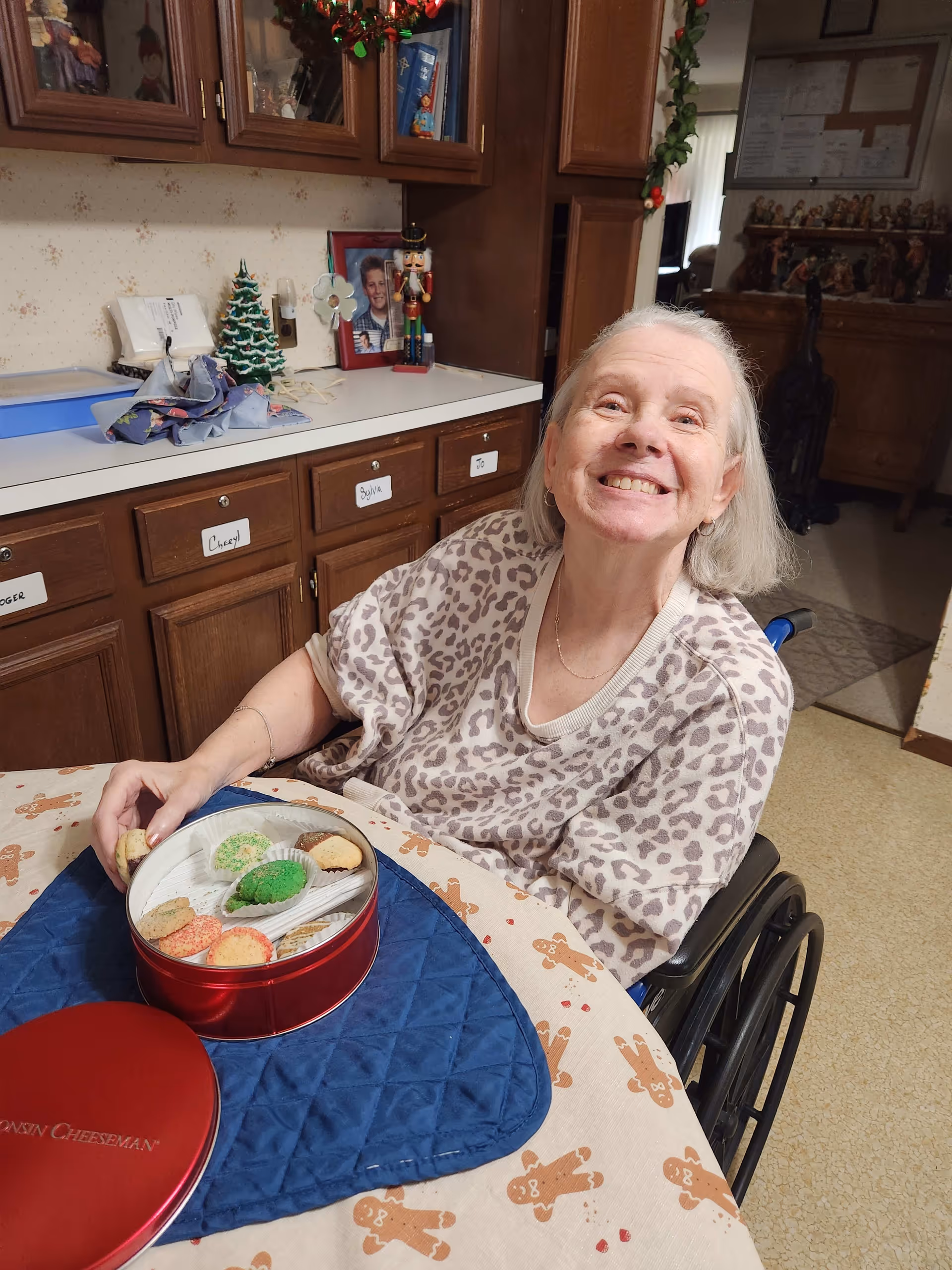 An elderly woman in a wheelchair smiling at the camera while sitting at a table with a gingerbread man patterned tablecloth. On the table is a red tin container with assorted cookies. Behind her are wooden cabinets with labels and holiday decorations including a small Christmas tree and a nutcracker figure.