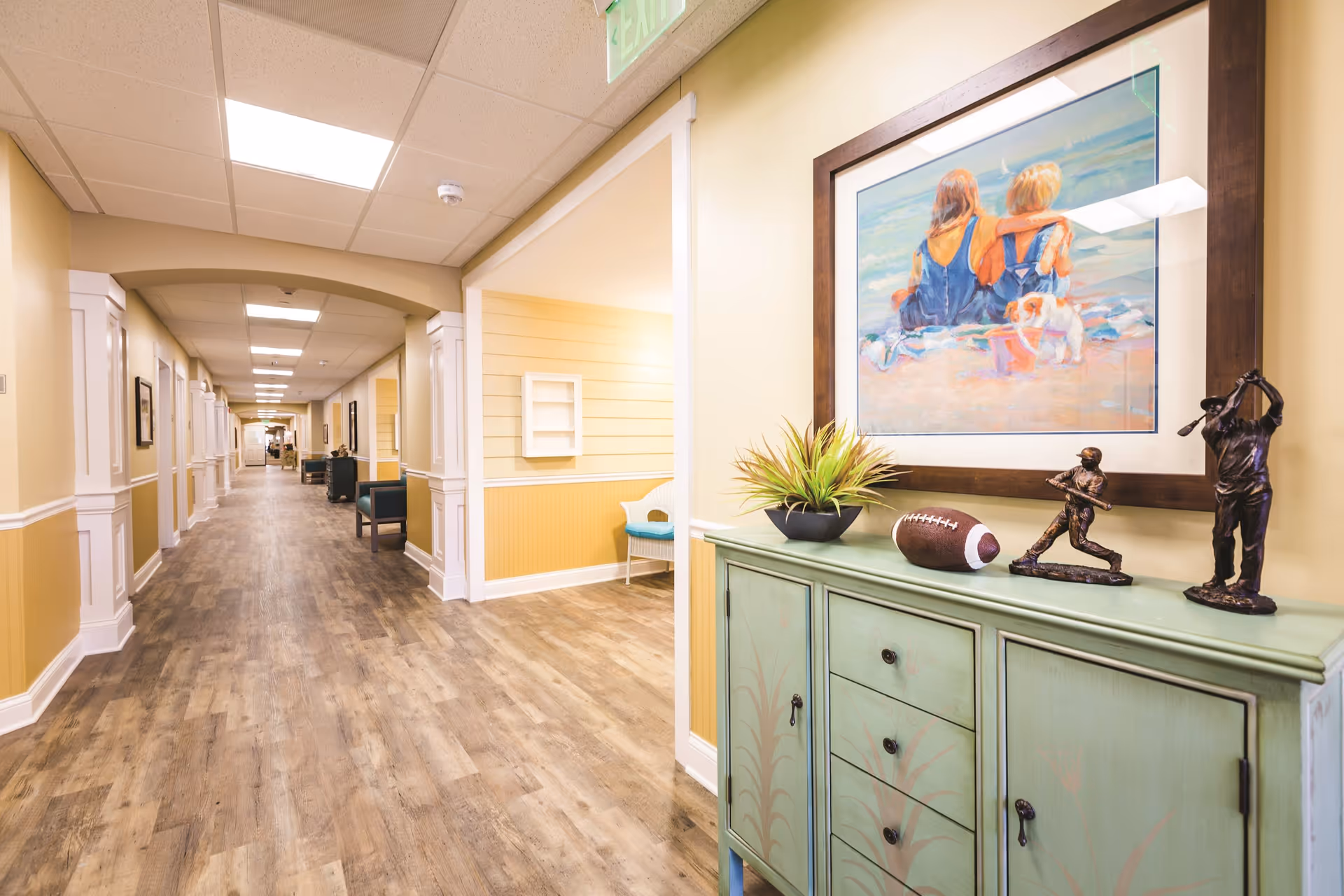 A long, well-lit hallway in a senior living facility with wood-style flooring and yellow walls. On the right side, there is a green cabinet with decorative items including a plant, a football, and two bronze statues of golfers. Above the cabinet hangs a framed painting of two children sitting on a beach with a small dog.