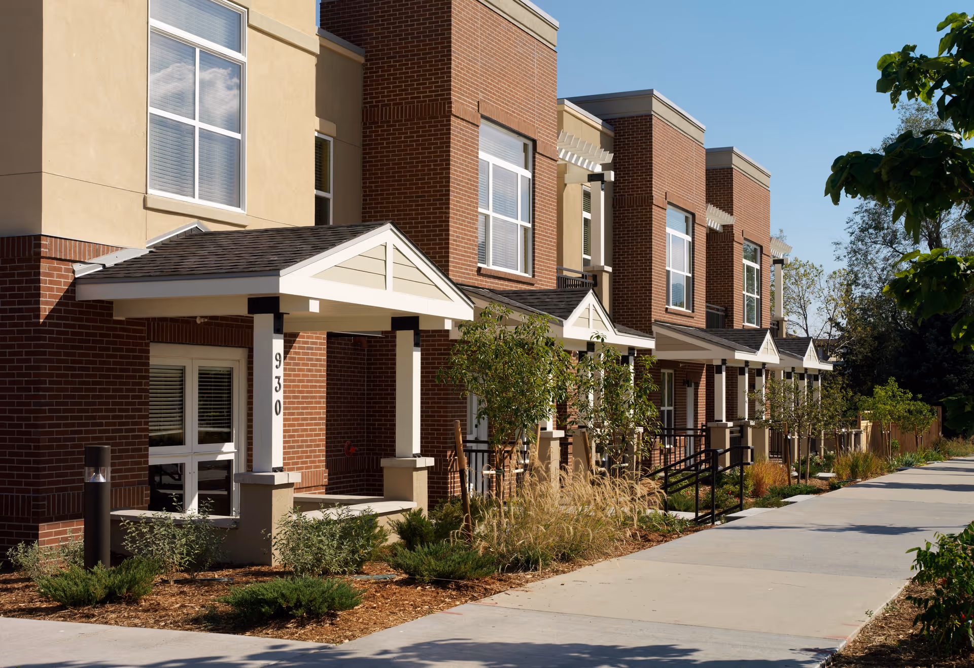 Front exterior of a brick and stucco senior living building with covered entrances, landscaping, and a sidewalk.