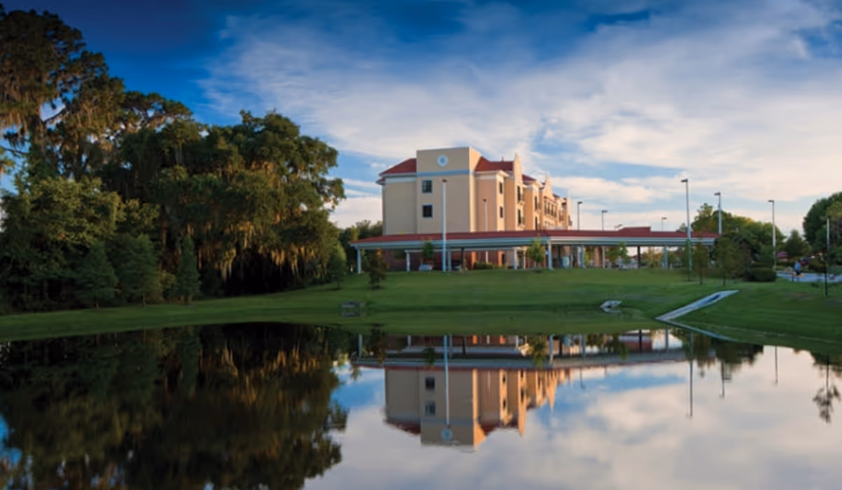 A multi-story senior living building with a covered entrance reflected in a pond under a blue sky.