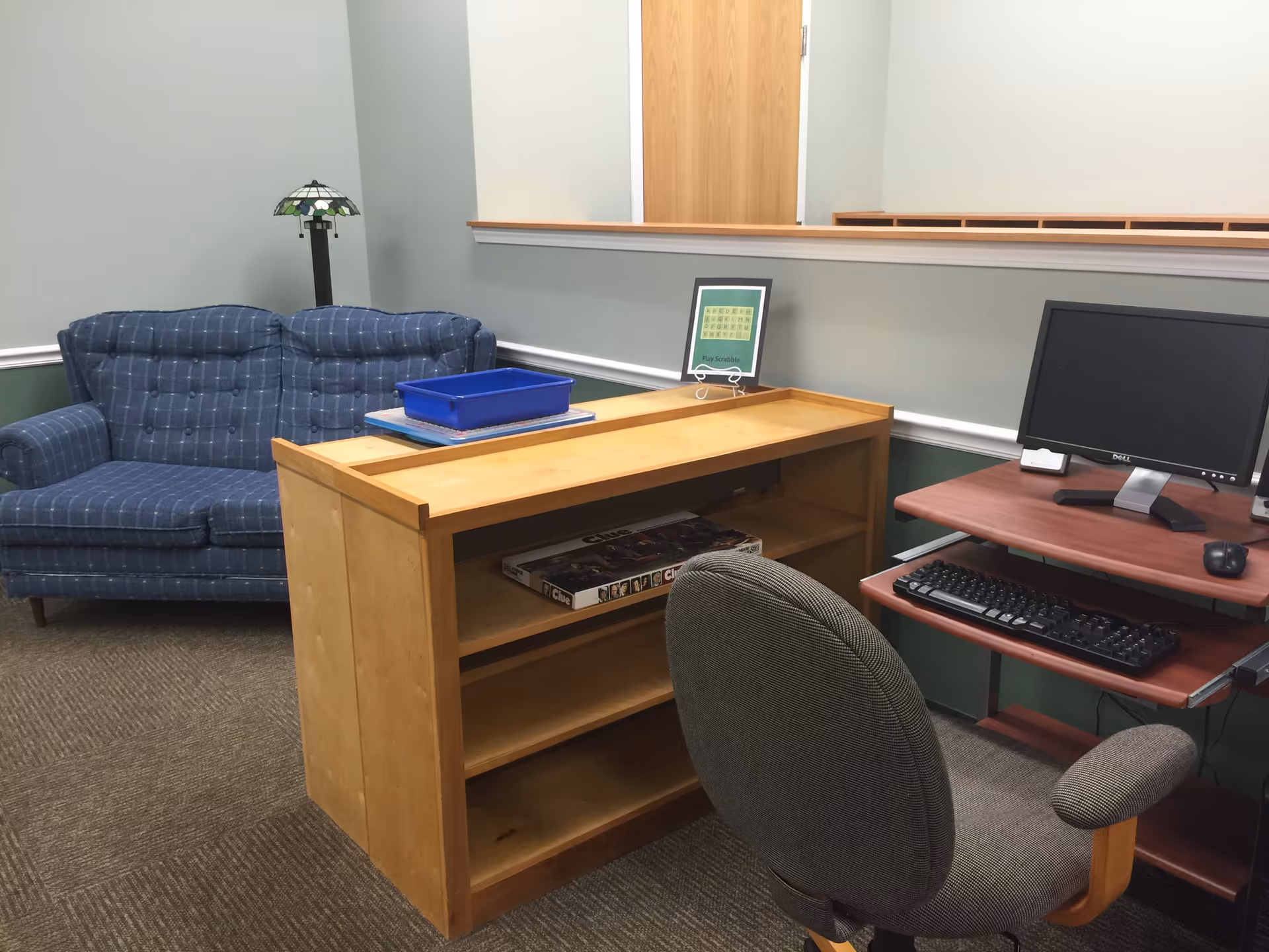Interior room with a blue patterned loveseat, a wooden shelving unit with a board game on the middle shelf, a framed sign that says Play Scrabble, a computer desk with a monitor, keyboard, and mouse, and an office chair.