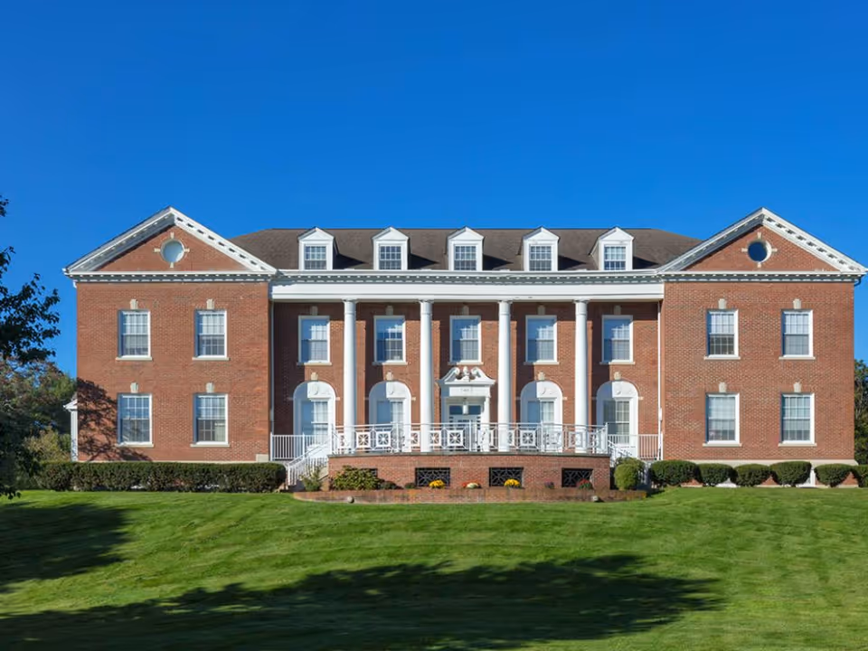 Brick neoclassical-style building with white columns sits on a green lawn under a blue sky.