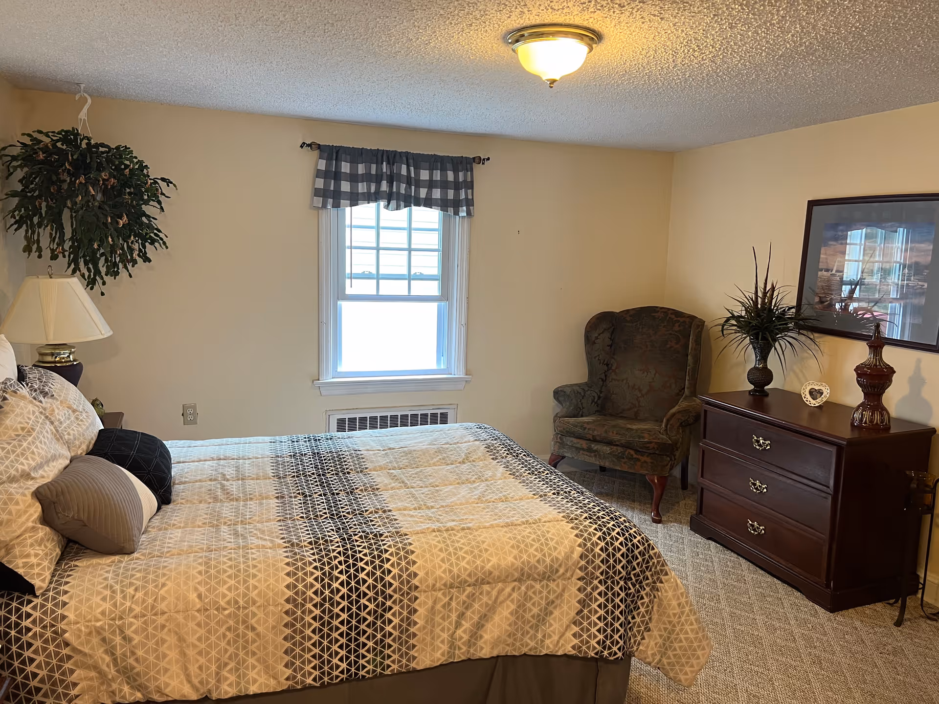 A cozy bedroom with a bed covered in a patterned quilt and several pillows. There is a window with a checkered valance above a heating unit. To the right, there is a dark wooden dresser with decorative items and a framed picture hanging above it. A green upholstered armchair is placed in the corner next to the dresser. A hanging plant and a table lamp are visible on the left side of the bed.