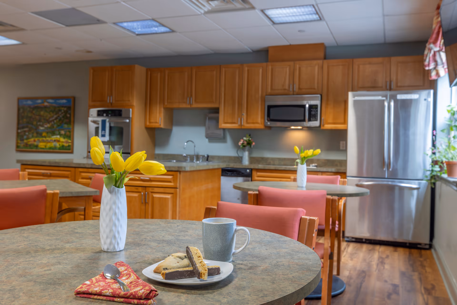 A bright kitchen and dining area with wooden cabinets, stainless steel appliances including a refrigerator, microwave, and oven. There are round tables with pink cushioned chairs, each table decorated with a white vase holding yellow tulips. On the closest table, there is a plate with biscotti and cookies, a gray mug, and a red patterned napkin with a spoon. A window with plants on the sill lets in natural light.