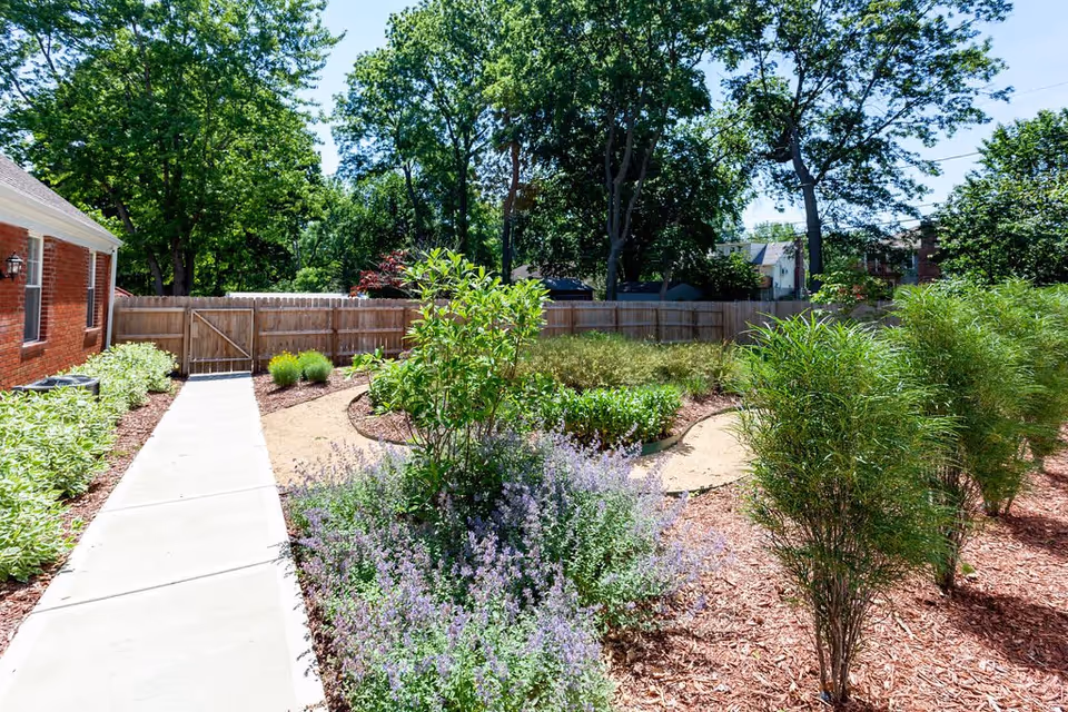 A sunny outdoor garden area at Golden Oaks Memory Care Home featuring a concrete walkway, various green shrubs and plants, a wooden fence, and tall trees in the background under a clear blue sky.