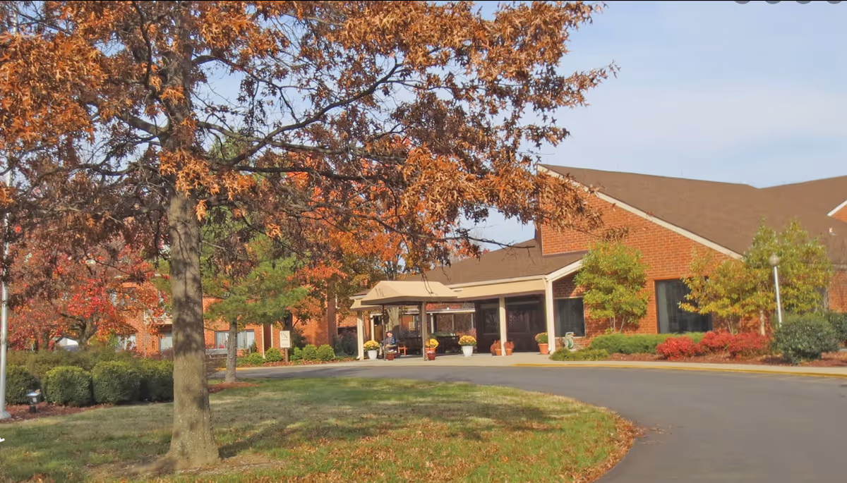 Exterior view of Westminster Village Kentuckiana, showing a brick building with a covered entrance. The scene includes a curved driveway, green bushes, trees with autumn-colored leaves, and a clear sky.