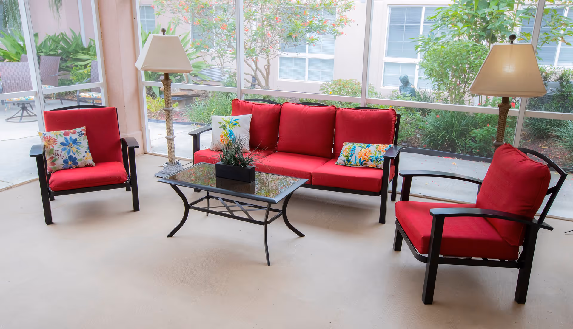 Sunroom seating area with red-cushioned sofa and chairs, a glass coffee table and lamps in front of large windows overlooking a garden.