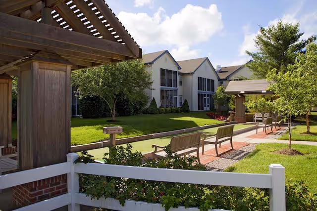 Sunlit courtyard with benches, pergolas, a brick walkway, and a multi-story residential building in the background.