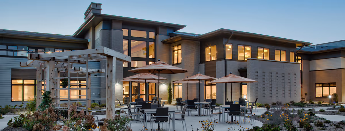 Outdoor patio area of a senior living facility with several tables and chairs under large umbrellas, surrounded by landscaped garden beds and a wooden pergola structure, with a modern two-story building in the background illuminated by warm interior lights at dusk.