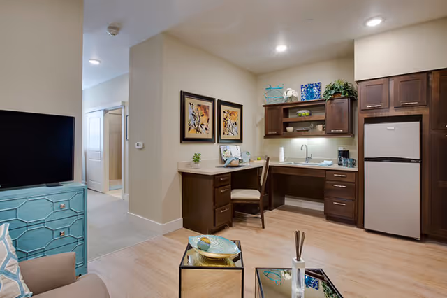Open studio living room and kitchenette with dark wood cabinets, a small desk area, stainless refrigerator, TV, and coffee table.