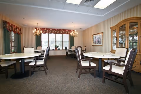 A dining room with round tables and cushioned chairs arranged neatly. The room features striped wallpaper, two chandeliers, a large window with floral valances and green curtains, and a wooden china cabinet displaying glassware and plates. A framed painting hangs on the wall.