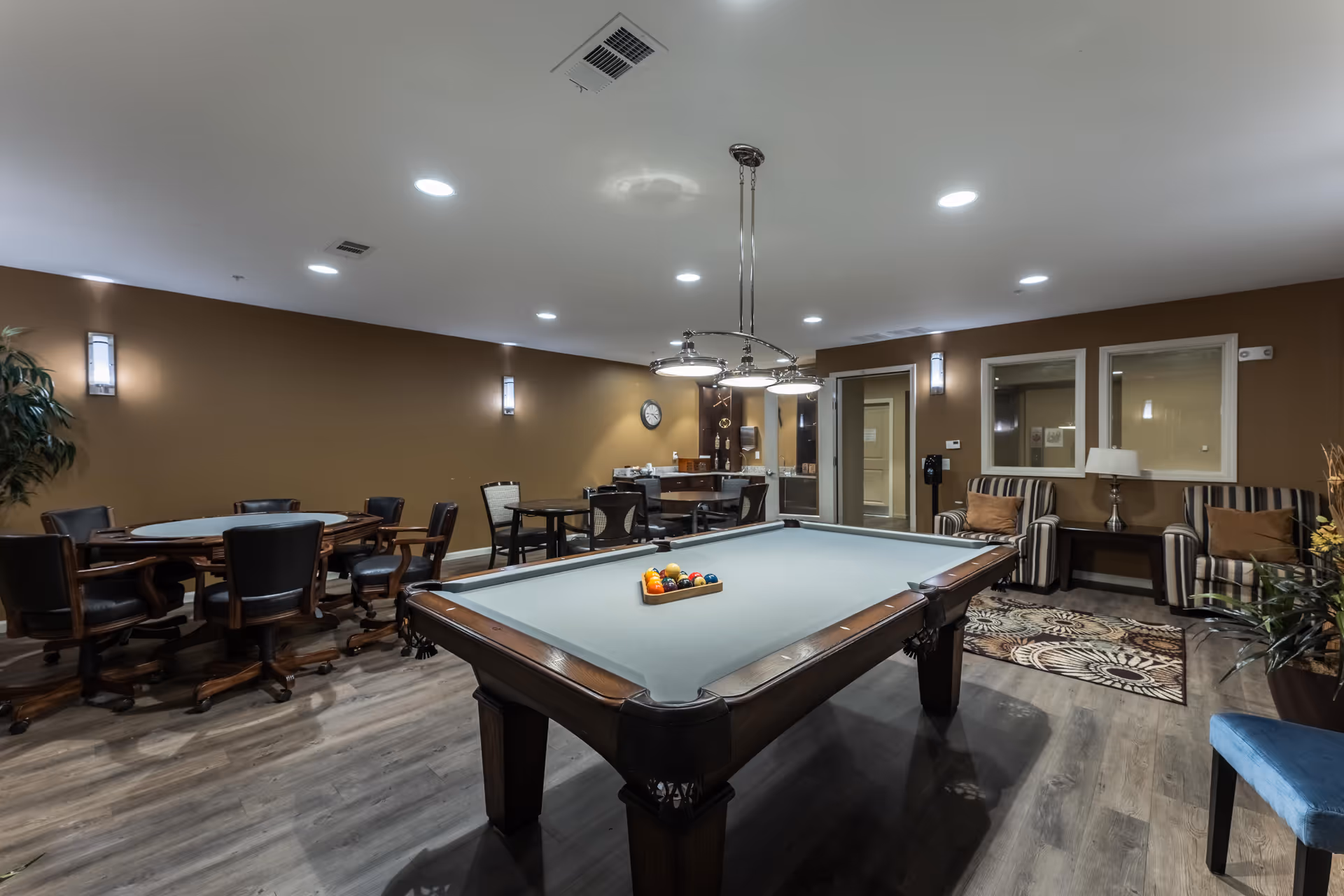 A recreational room with a pool table in the center, surrounded by several chairs and tables. The room has brown walls, wood flooring, and modern ceiling lights. There are two striped armchairs with a small table and lamp between them, a patterned rug, and a plant in the corner.