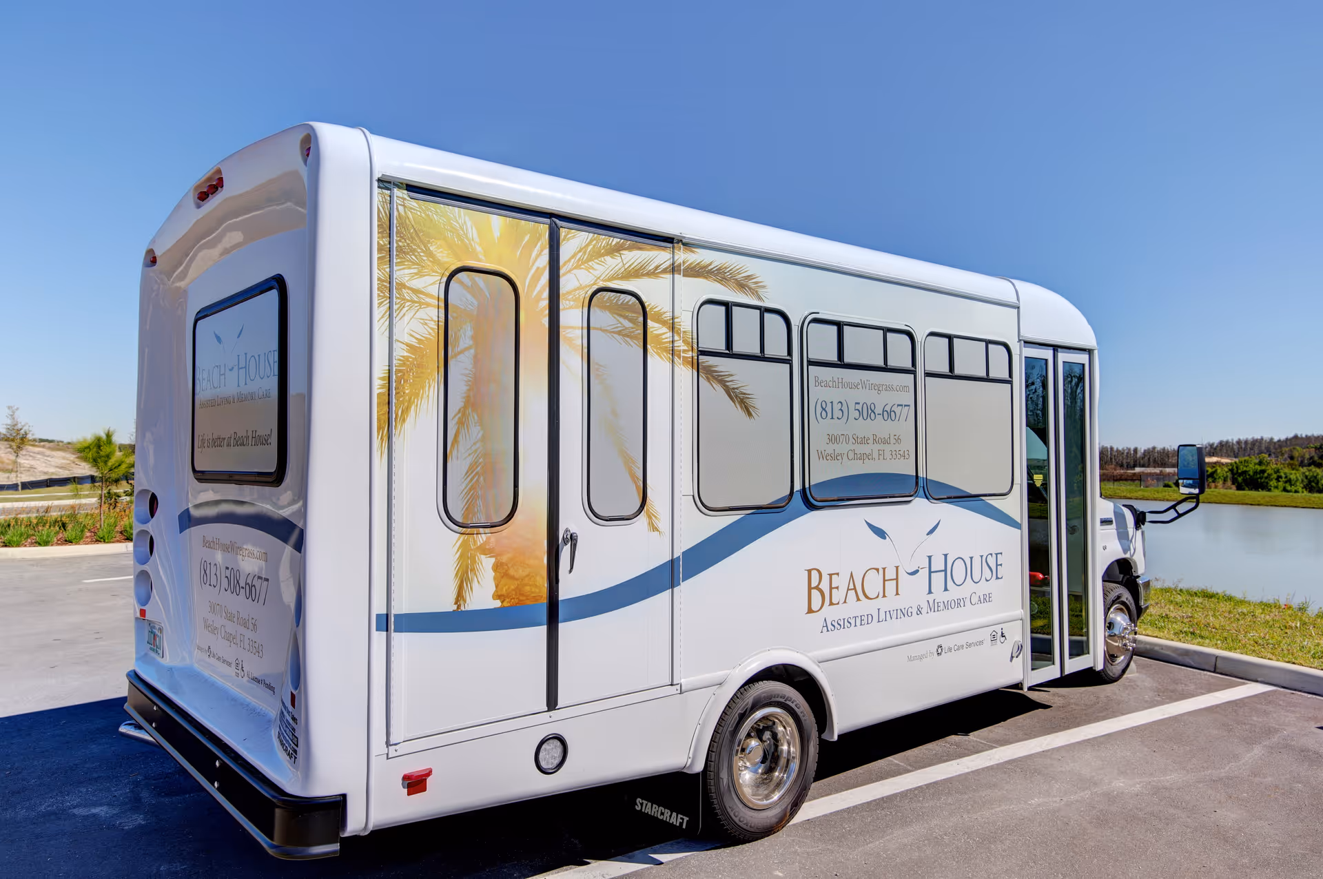A white shuttle bus parked outdoors near a body of water, branded with Beach House Assisted Living & Memory Care logos and contact information, featuring a palm tree graphic on the side.
