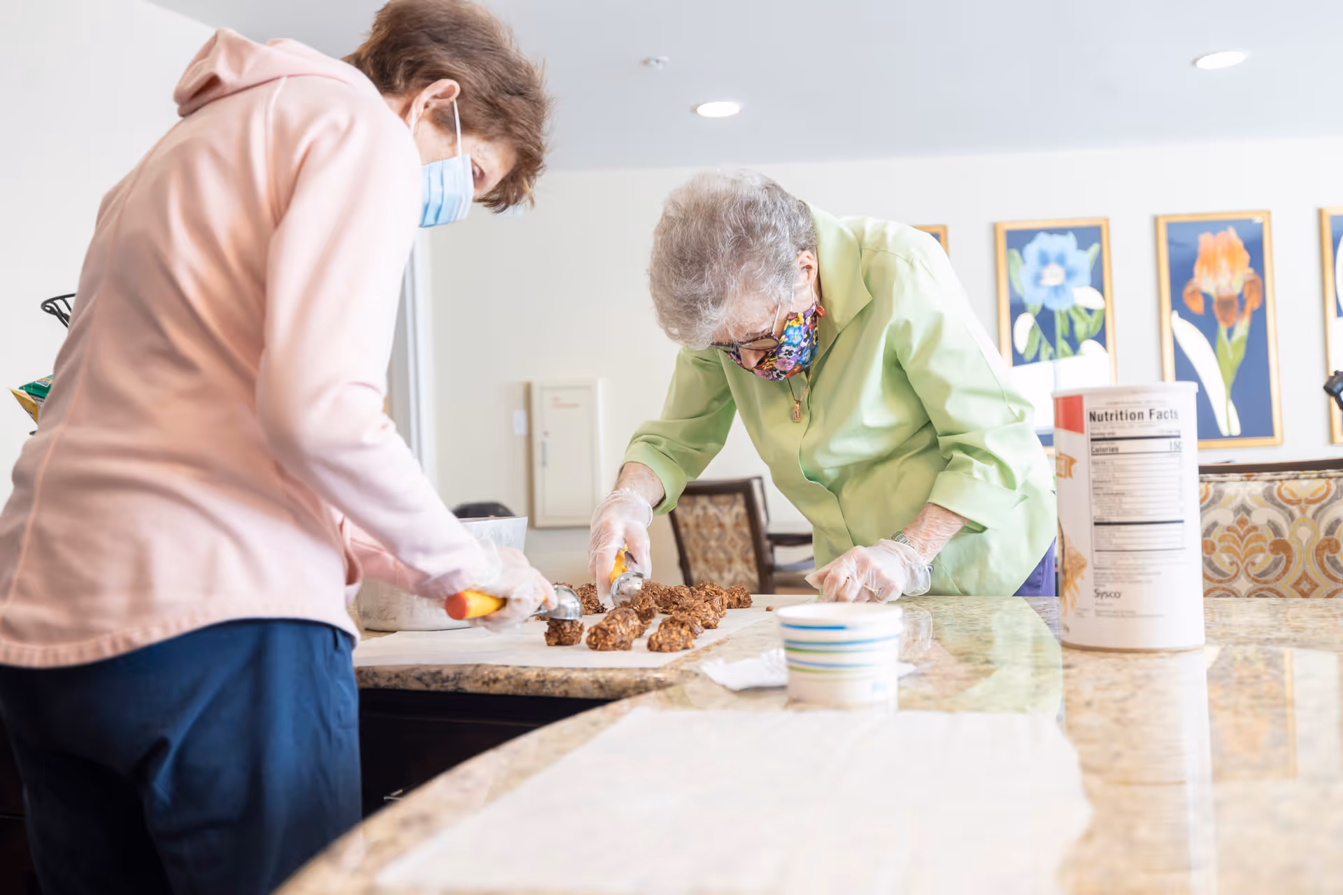 Two elderly women wearing face masks and gloves are scooping cookie dough onto a countertop in a well-lit room with floral artwork on the wall.