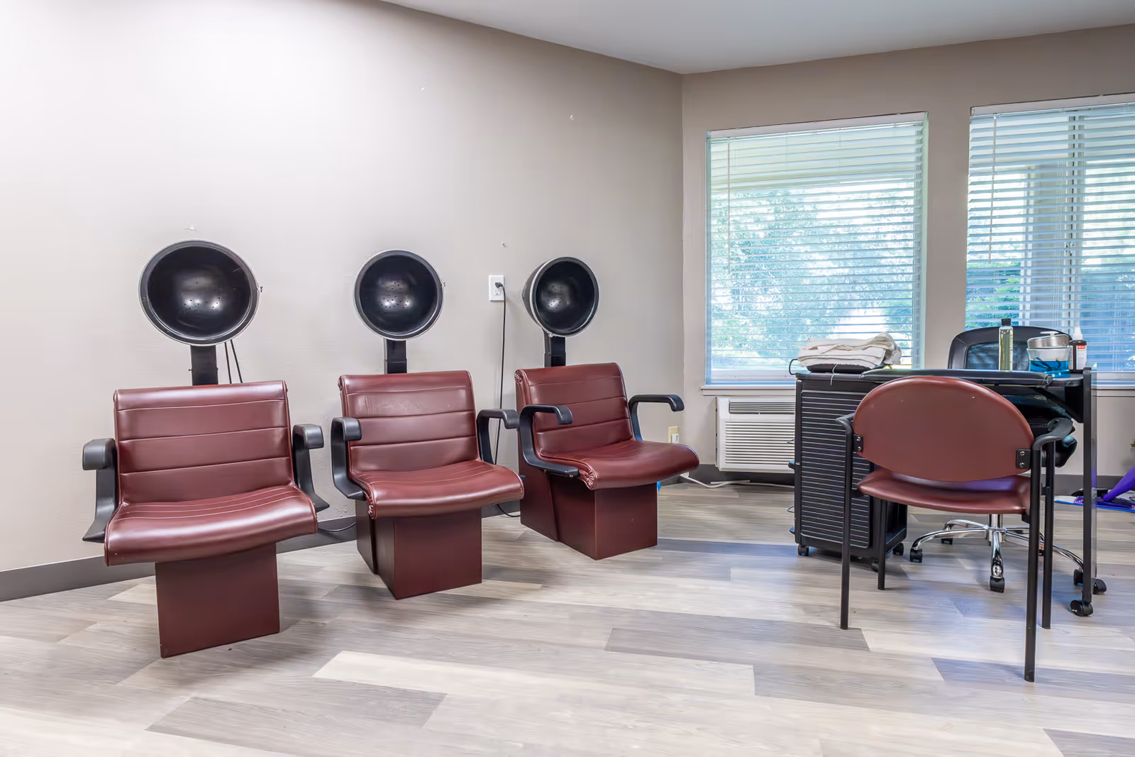 Three maroon salon chairs with hooded hair dryers along a wall and a small workstation by windows.