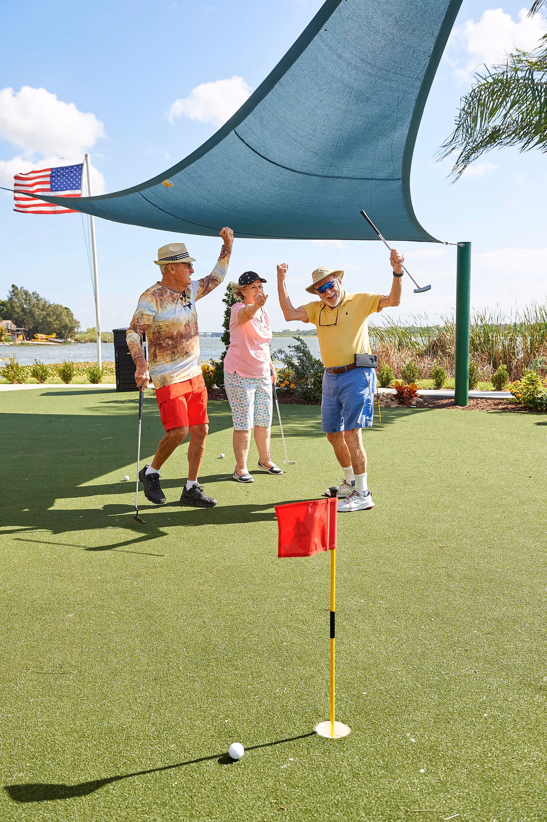Three elderly people playing mini golf on a sunny day near a lake, with a red flag marking the hole and an American flag in the background. They appear to be celebrating a successful shot under a blue shade sail.