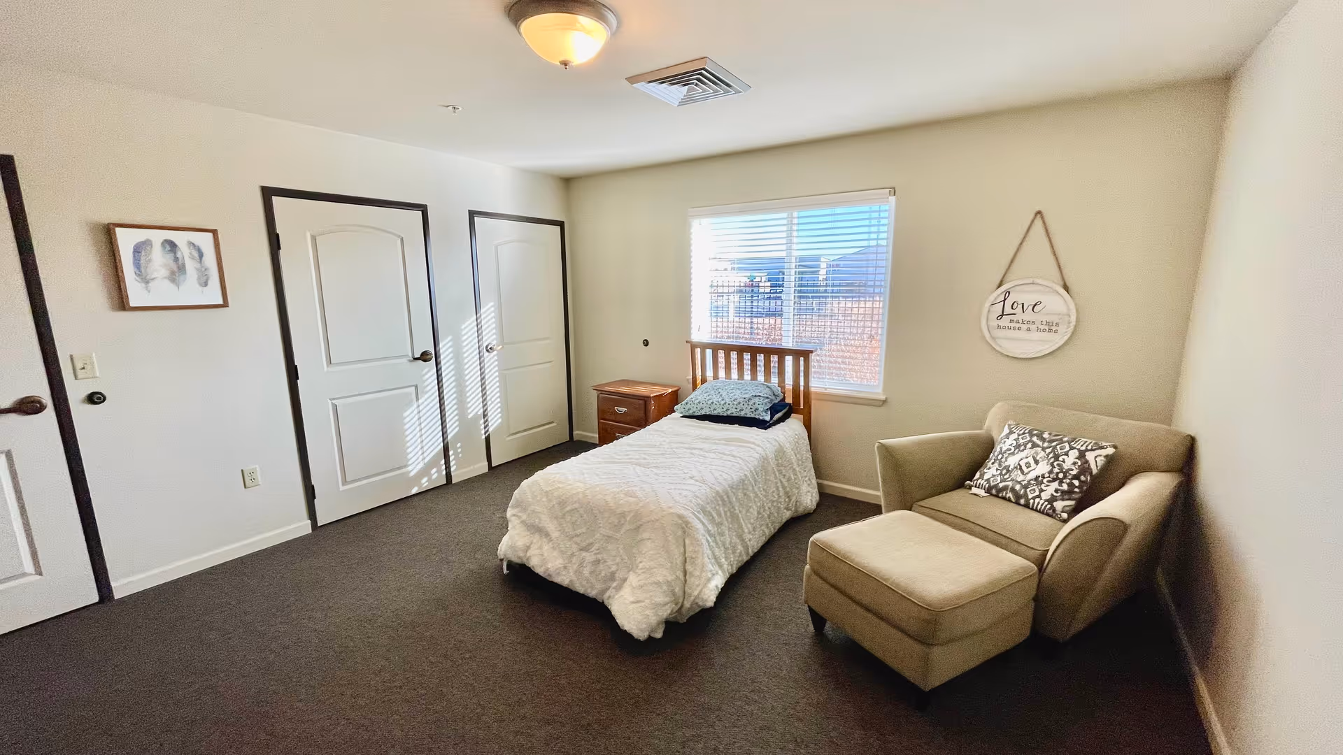 A simple bedroom with a single bed covered in a white comforter, a wooden headboard, and a small wooden nightstand next to it. There is a beige armchair with a matching ottoman and a patterned pillow on the right side near the window. The window has white blinds, and a decorative sign hangs on the wall that reads 'Love makes this house a home.' The room has three white doors with dark trim and a framed picture on the left wall. The floor is carpeted in a dark color.