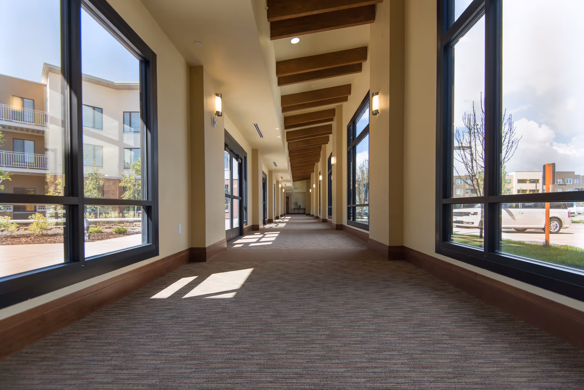 Sunlit corridor with large floor-to-ceiling windows, exposed wooden ceiling beams, and a carpeted floor looking out onto the courtyard.