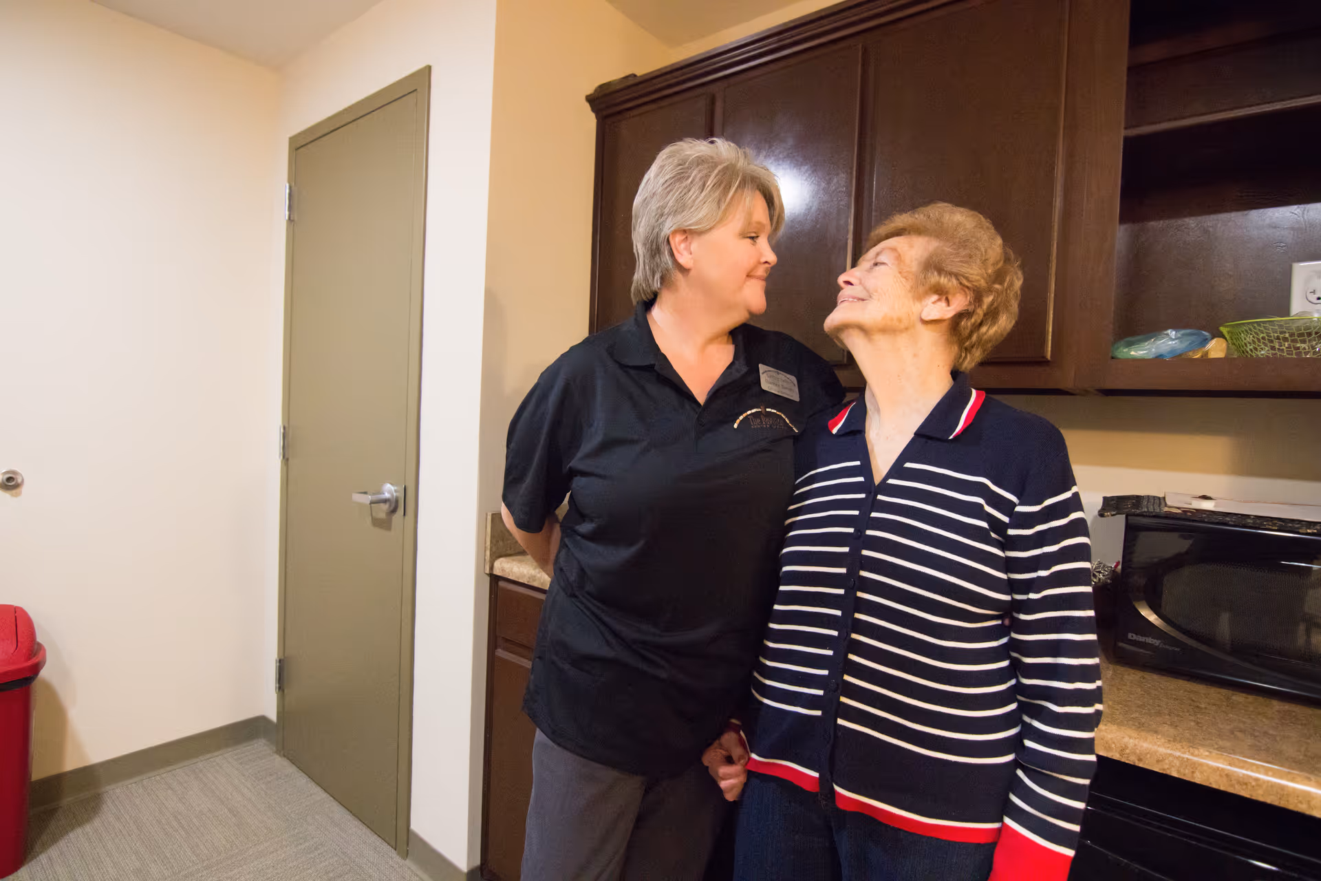 A caregiver and an elderly woman standing close together in a kitchen area, smiling and looking at each other. The kitchen has dark wooden cabinets, a microwave, and a countertop. There is a closed door and a red trash bin in the background.