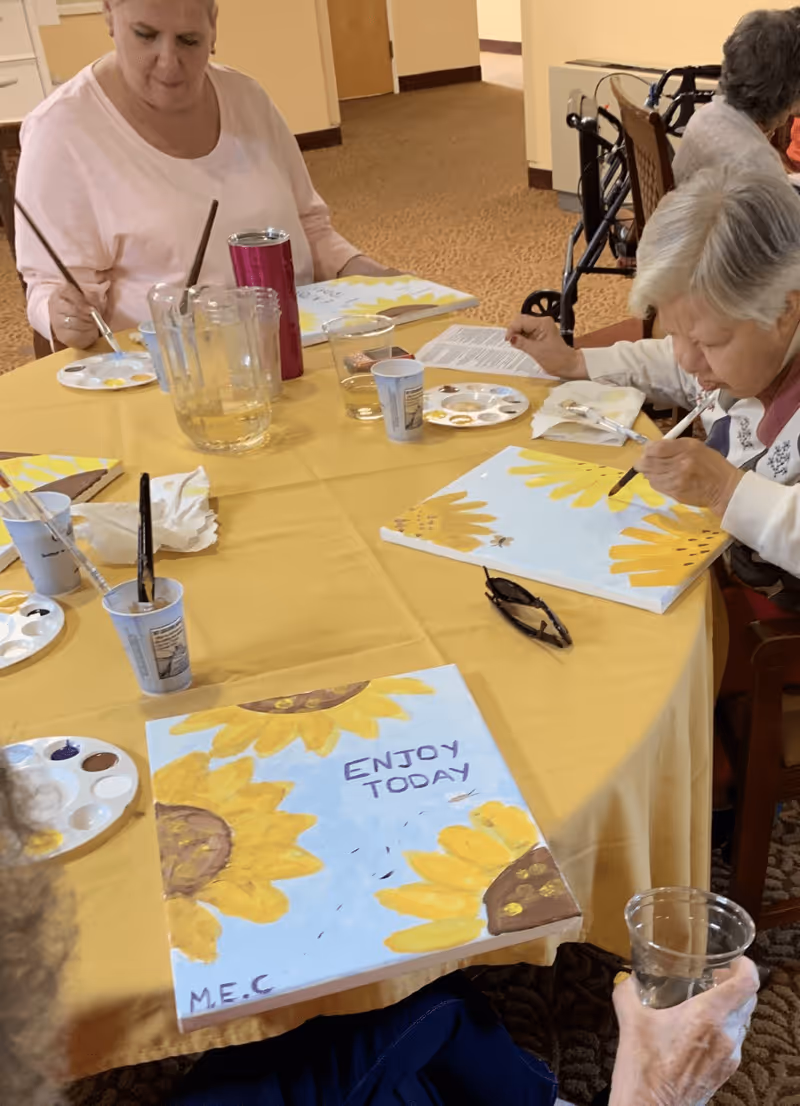 Several elderly individuals seated around a table covered with a yellow tablecloth, engaged in painting sunflower-themed canvases. One canvas has the words 'ENJOY TODAY' painted on it. Various painting supplies, cups, and water pitchers are on the table.