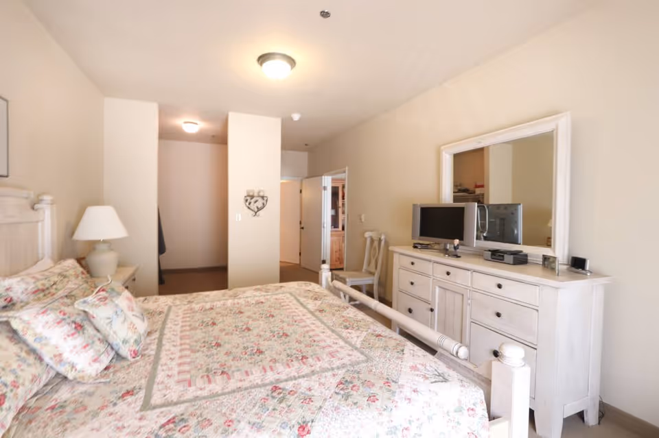 A bedroom with a floral patterned bedspread and pillows on a white wooden bed frame. There is a white dresser with a mirror above it, a small TV, and a chair next to the dresser. The walls are painted beige and there is a ceiling light fixture. The room has an open doorway leading to another area.