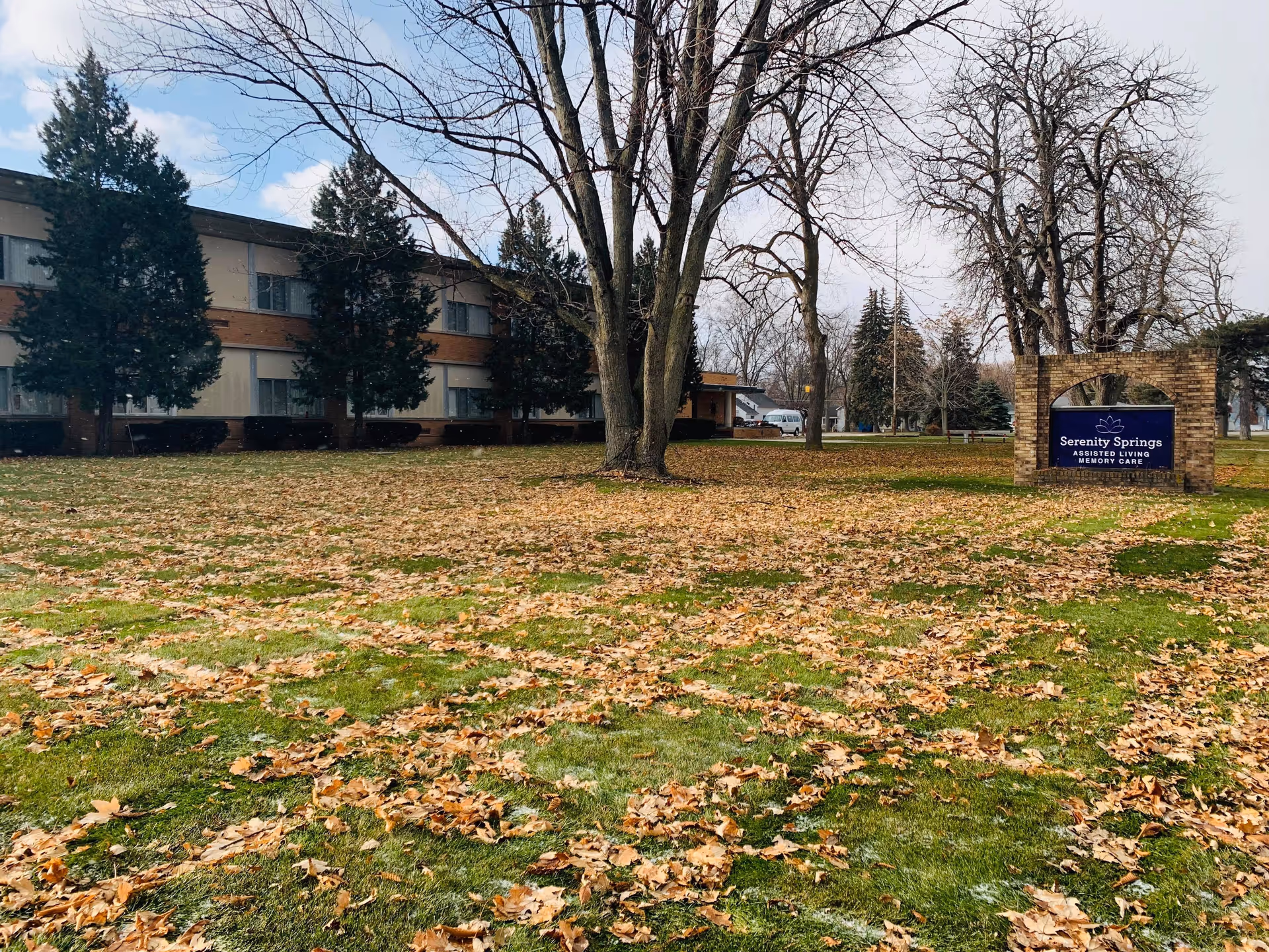 Exterior view of Serenity Springs assisted living and memory care facility with a lawn covered in fallen leaves, several leafless trees, and a brick sign displaying the facility's name.