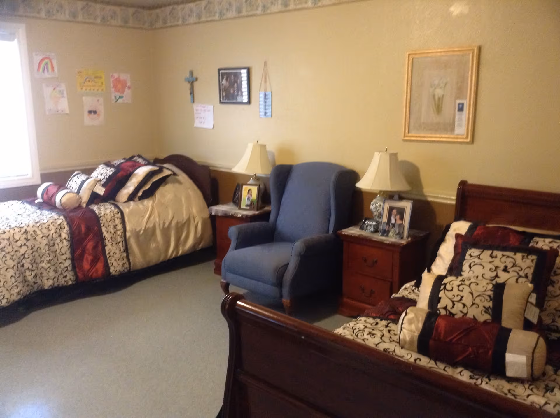 A cozy bedroom with two beds featuring matching patterned bedding in cream, black, and red colors. Between the beds is a blue upholstered armchair and two wooden nightstands with lamps and framed photos. The walls are decorated with a cross, framed pictures, and children's drawings near a window.