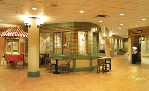 Interior view of a senior living facility with a central reception or information desk surrounded by chairs and tables. The area has tiled floors, beige walls, and a popcorn machine cart with a red and white striped canopy on the left. There is a hallway extending to the right with additional seating and lighting fixtures on the walls.