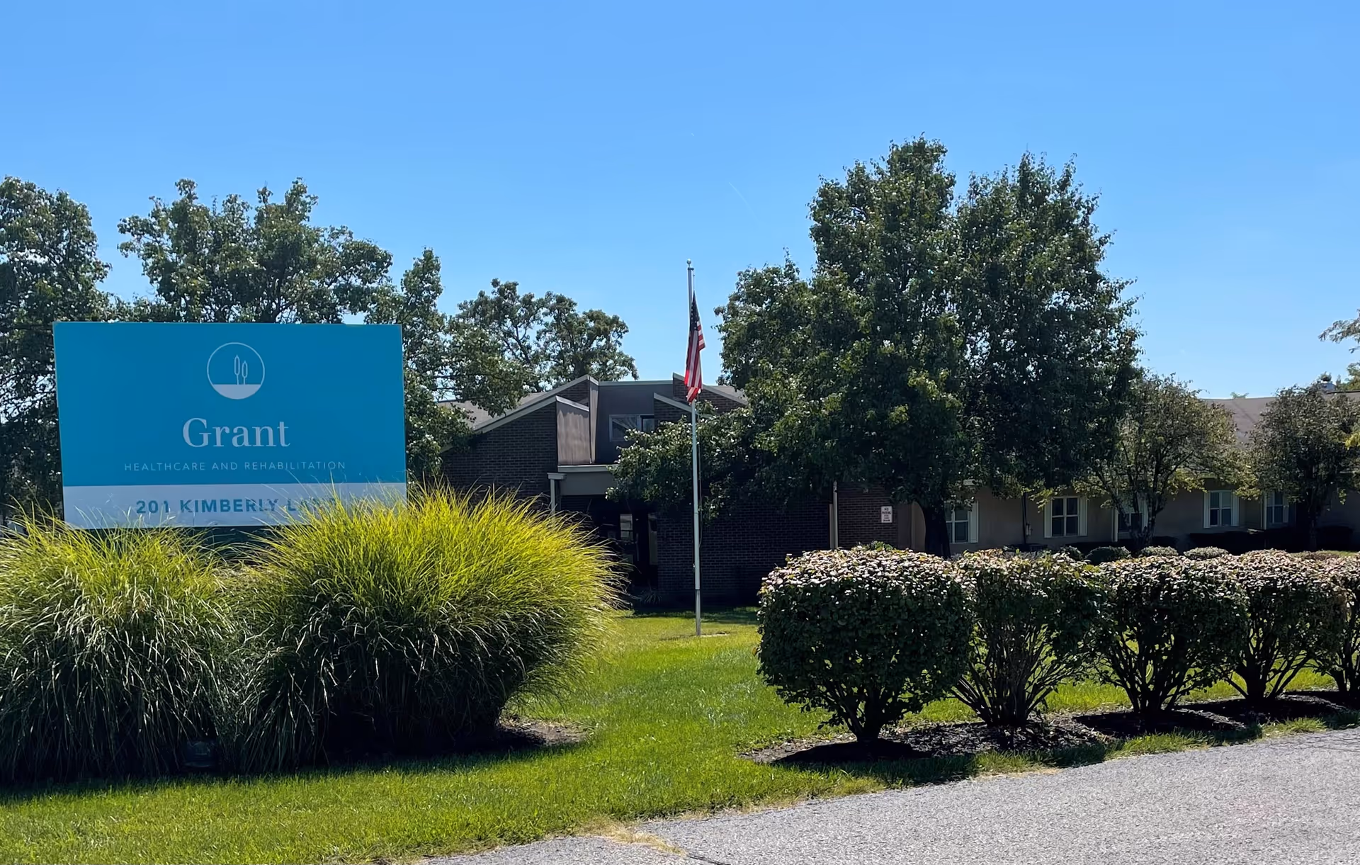 Exterior view of Grant Healthcare and Rehabilitation facility with a large blue sign displaying the facility name and address, surrounded by green bushes and trees under a clear blue sky.