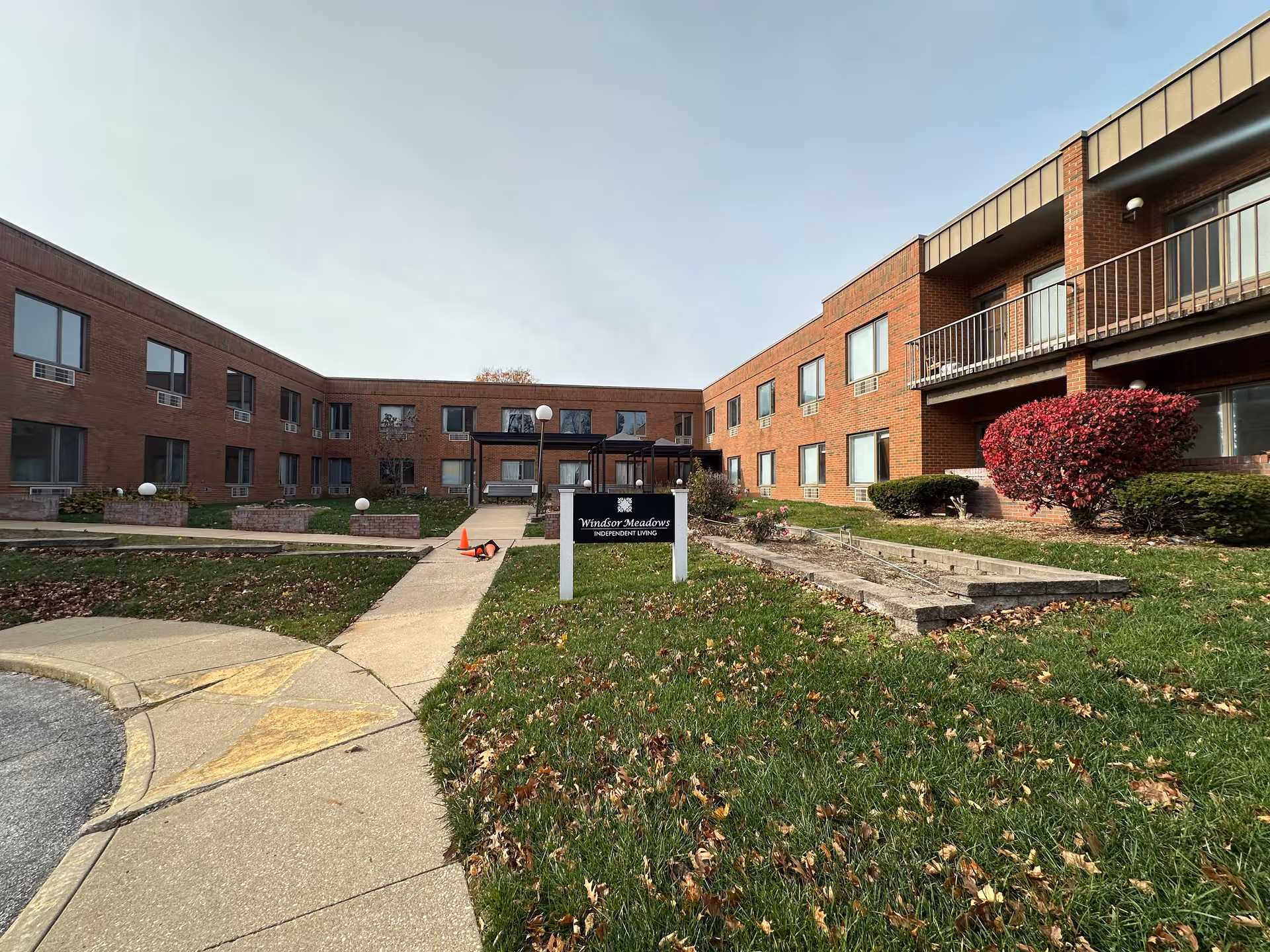 Front view of a two-story brick senior living building with a central entrance, walkway, lawn and a sign near the sidewalk.
