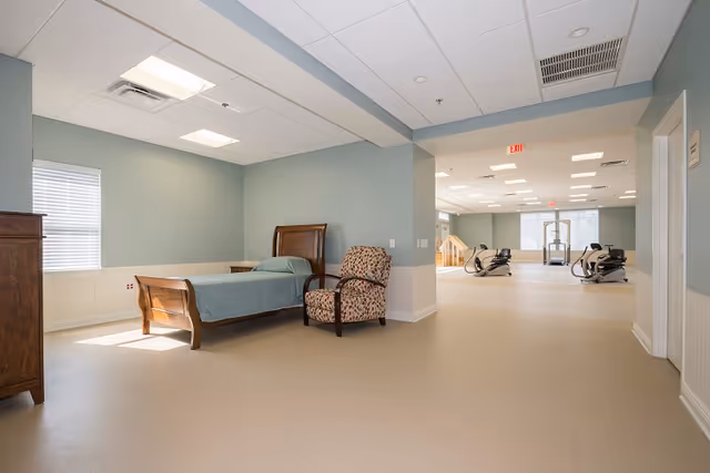 A room with a single wooden bed with a blue bedspread and a patterned armchair next to it. The walls are painted light blue with white wainscoting. There is a window with blinds on the left side. The room opens into a larger area with exercise equipment and large windows letting in natural light.