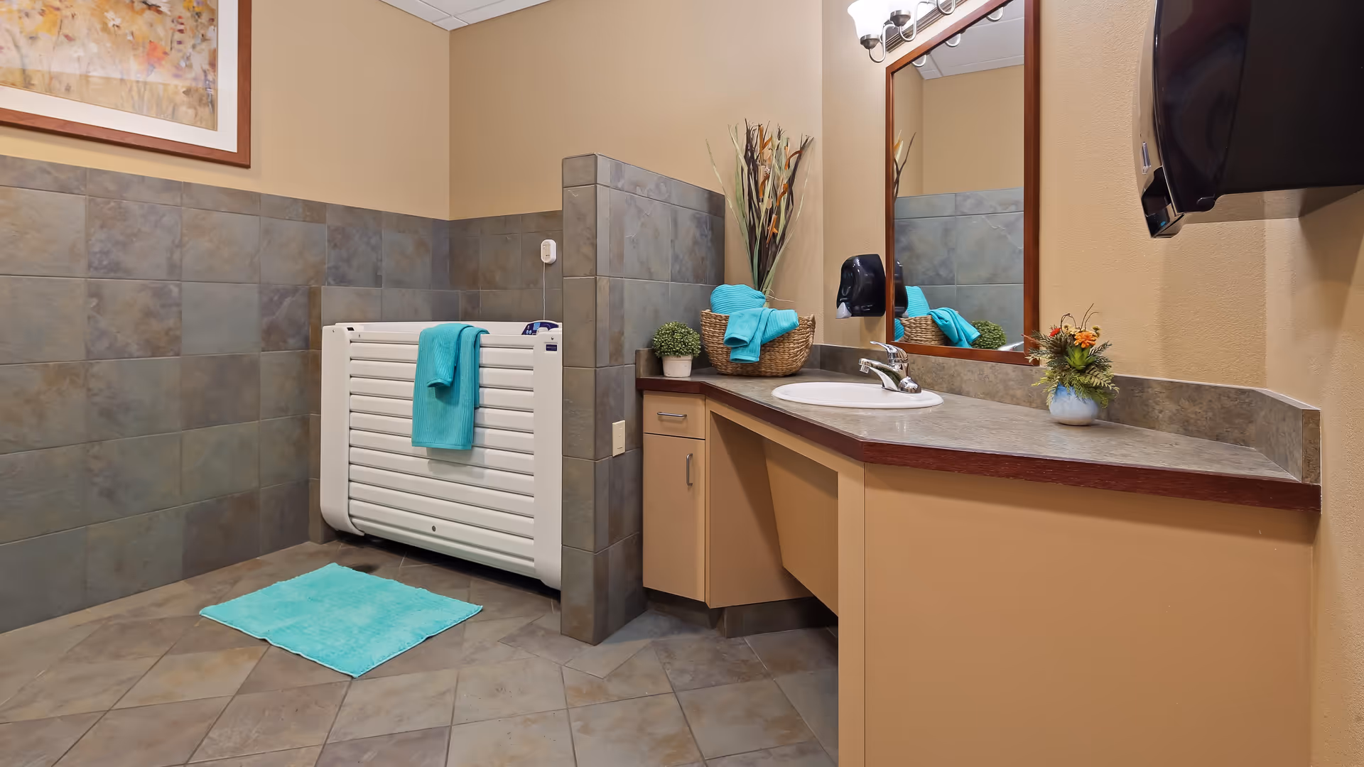 A clean bathroom with beige walls and tiled floor and lower walls. There is a white radiator with a turquoise towel hanging on it, a turquoise bath mat on the floor, a countertop with a sink, a large mirror above the sink, a black soap dispenser mounted on the wall, and decorative plants on the countertop. A framed artwork is partially visible on the upper left wall.