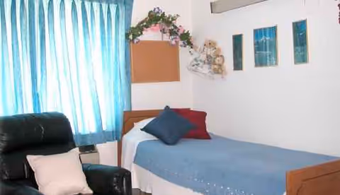 A bedroom in a senior living facility with a bed, blue curtains, and an armchair.