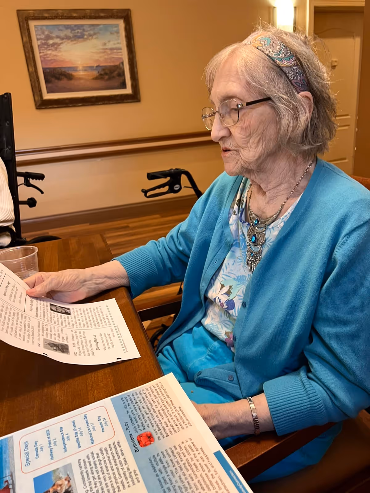 An elderly woman with glasses and a colorful headband is sitting at a wooden table in a room with warm lighting, reading printed papers. She is wearing a blue cardigan over a floral blouse and a statement necklace. In the background, there is a framed painting on the wall and a walker.