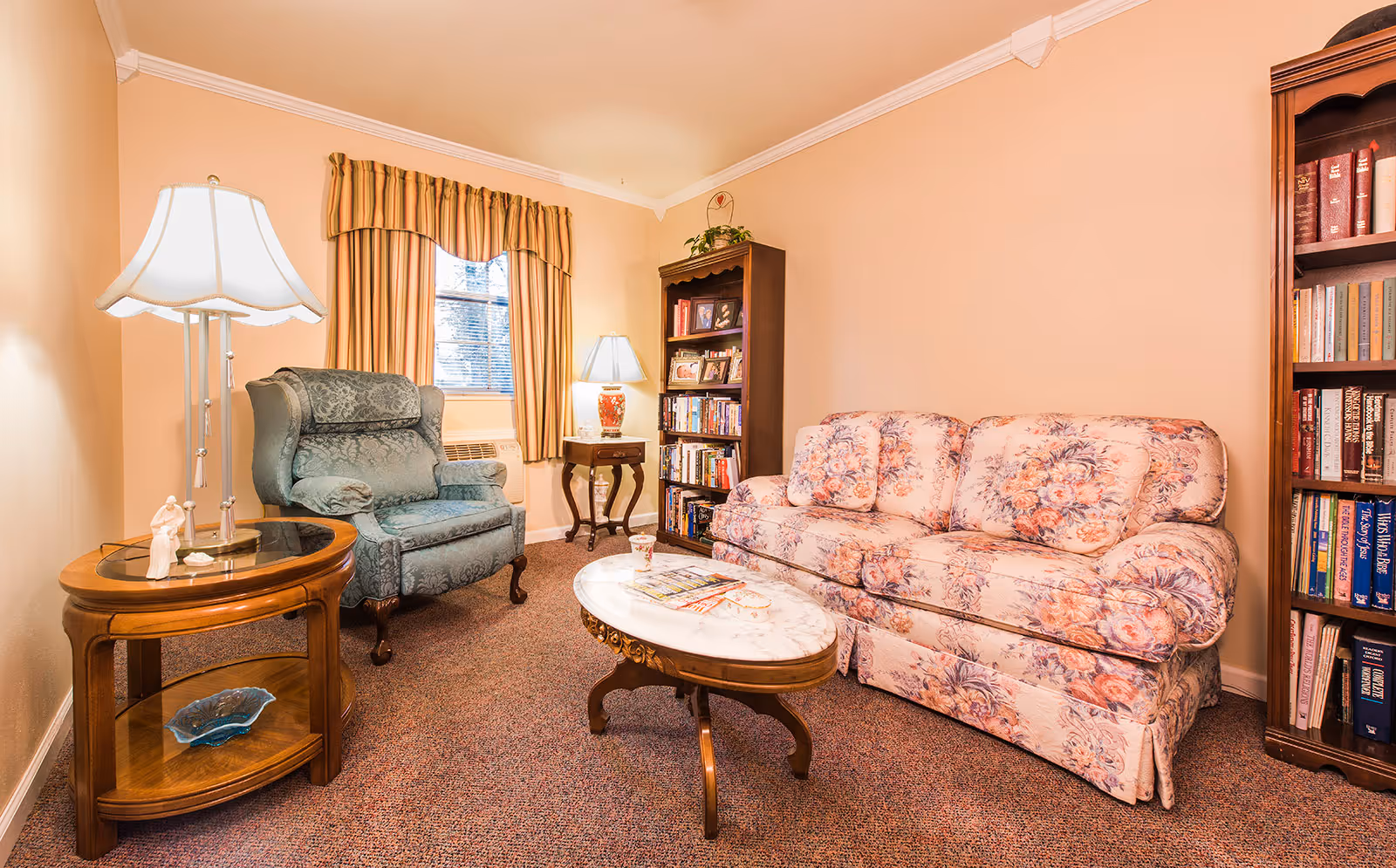 Cozy living room with a floral sofa, upholstered armchair, marble-top coffee table, side tables with lamps, bookshelves, and a curtained window.