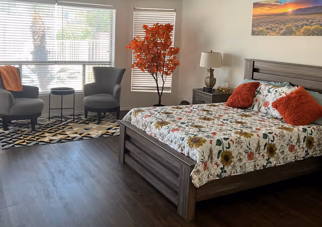 A cozy bedroom with a wooden bed frame and floral bedspread featuring sunflowers and other flowers. Two orange throw pillows are on the bed. Next to the bed is a nightstand with a lamp, clock, and small decorative items. Two gray armchairs with a small round table between them sit on a geometric patterned rug near large windows with blinds. A tall artificial tree with orange leaves is positioned between the windows. A landscape painting hangs on the wall above the bed.