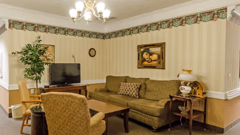 A cozy living room with a brown sofa, a patterned armchair, a wooden coffee table, and a TV on a stand. The walls have beige striped wallpaper with a decorative border near the ceiling. There is a framed painting, a clock, a potted plant, and a vintage-style lamp on a side table.