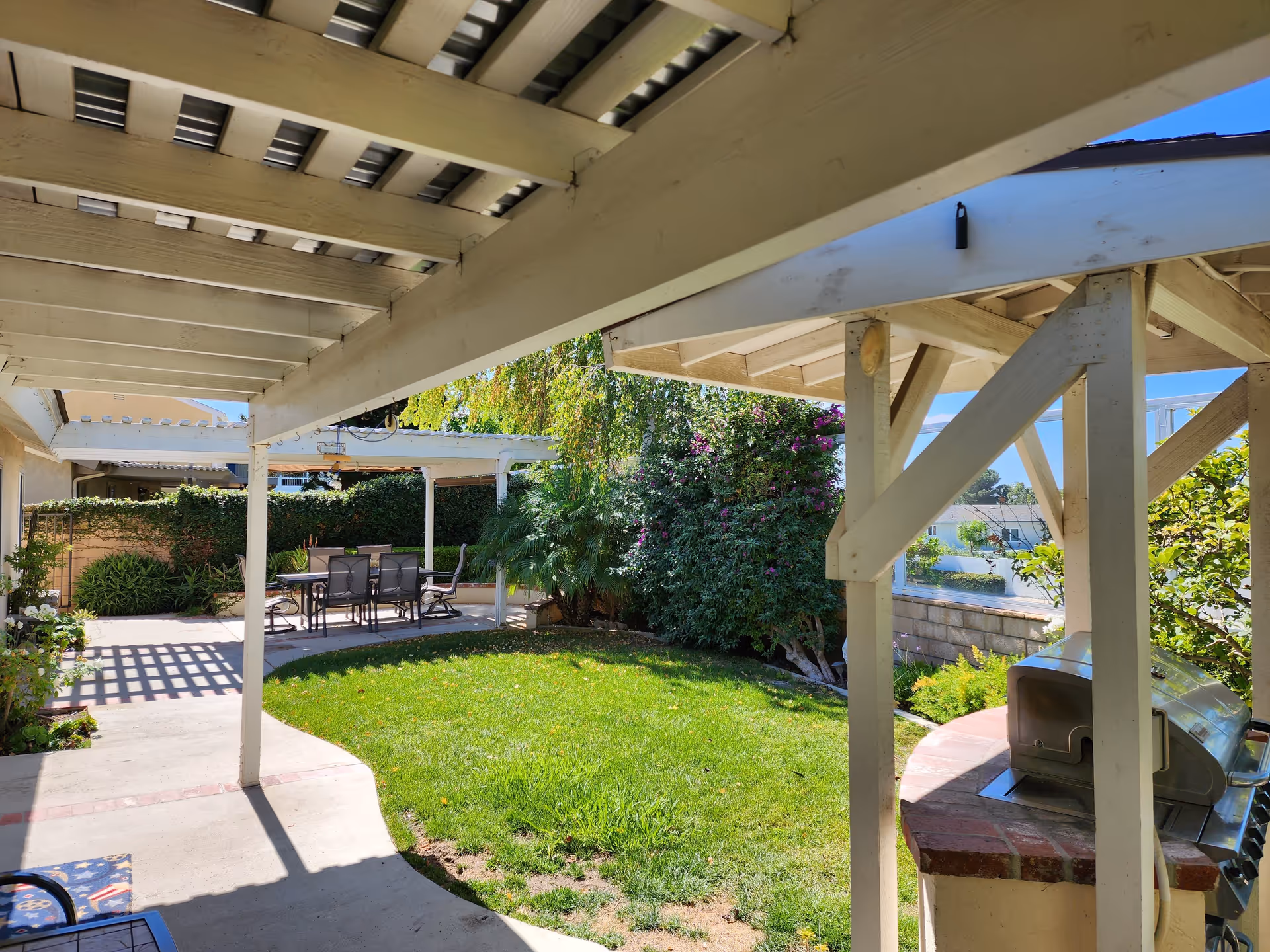 Outdoor patio area with a covered walkway, green lawn, and a dining table with chairs under a pergola. There are bushes and trees surrounding the space, and a built-in barbecue grill on the right side.