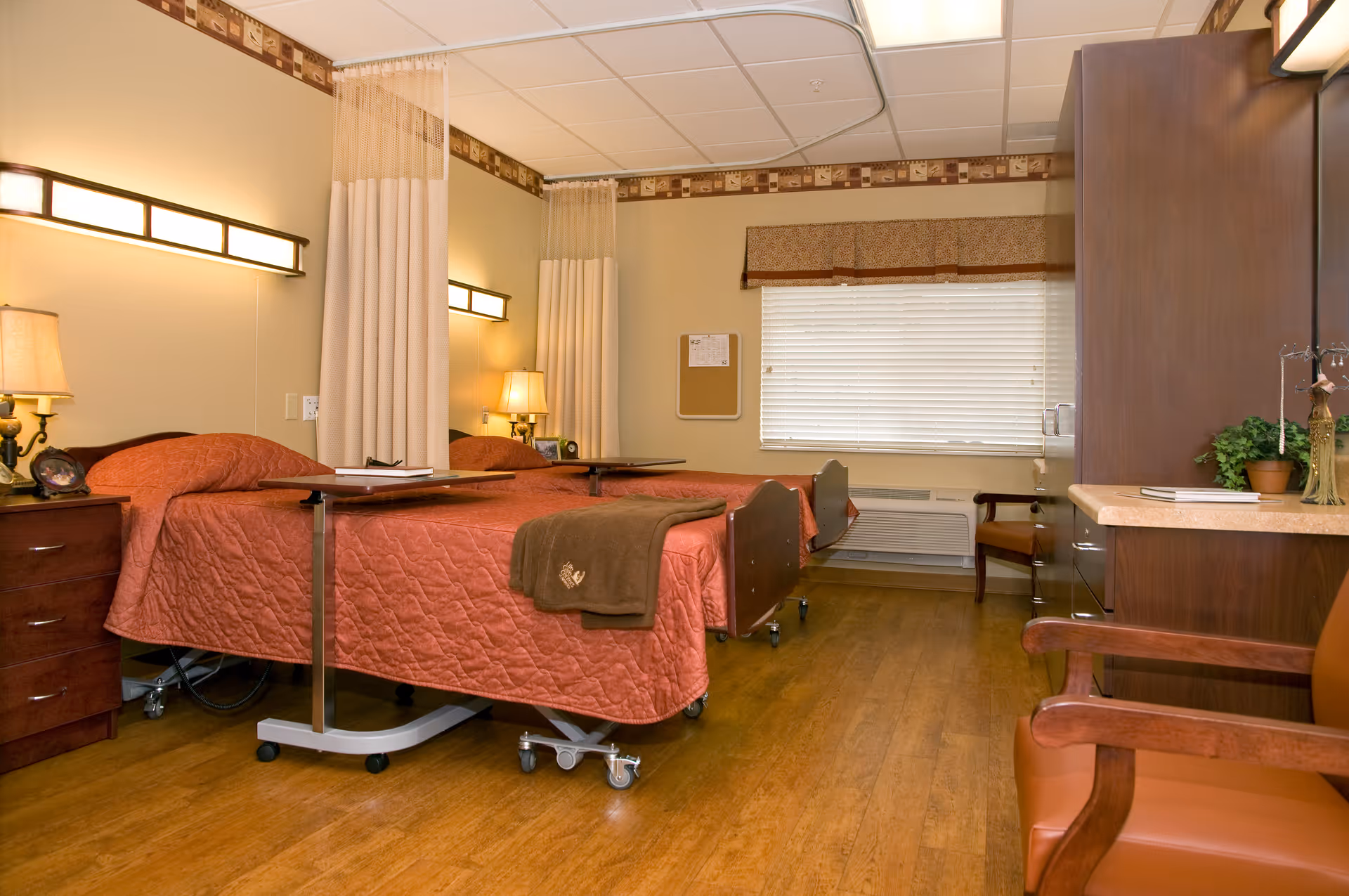 A tidy senior living bedroom with two hospital-style beds, bedside tables, chairs, and warm lighting.